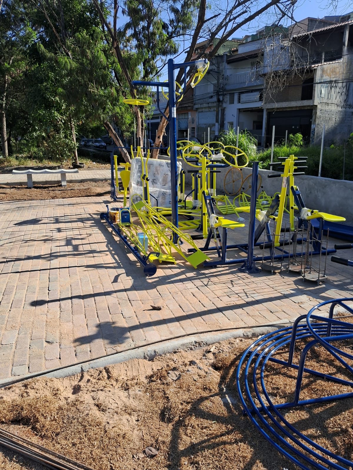 Espaço aberto cimentado, aparelhos de exercício nas cores amarelo e azul no canto prontos para instalação